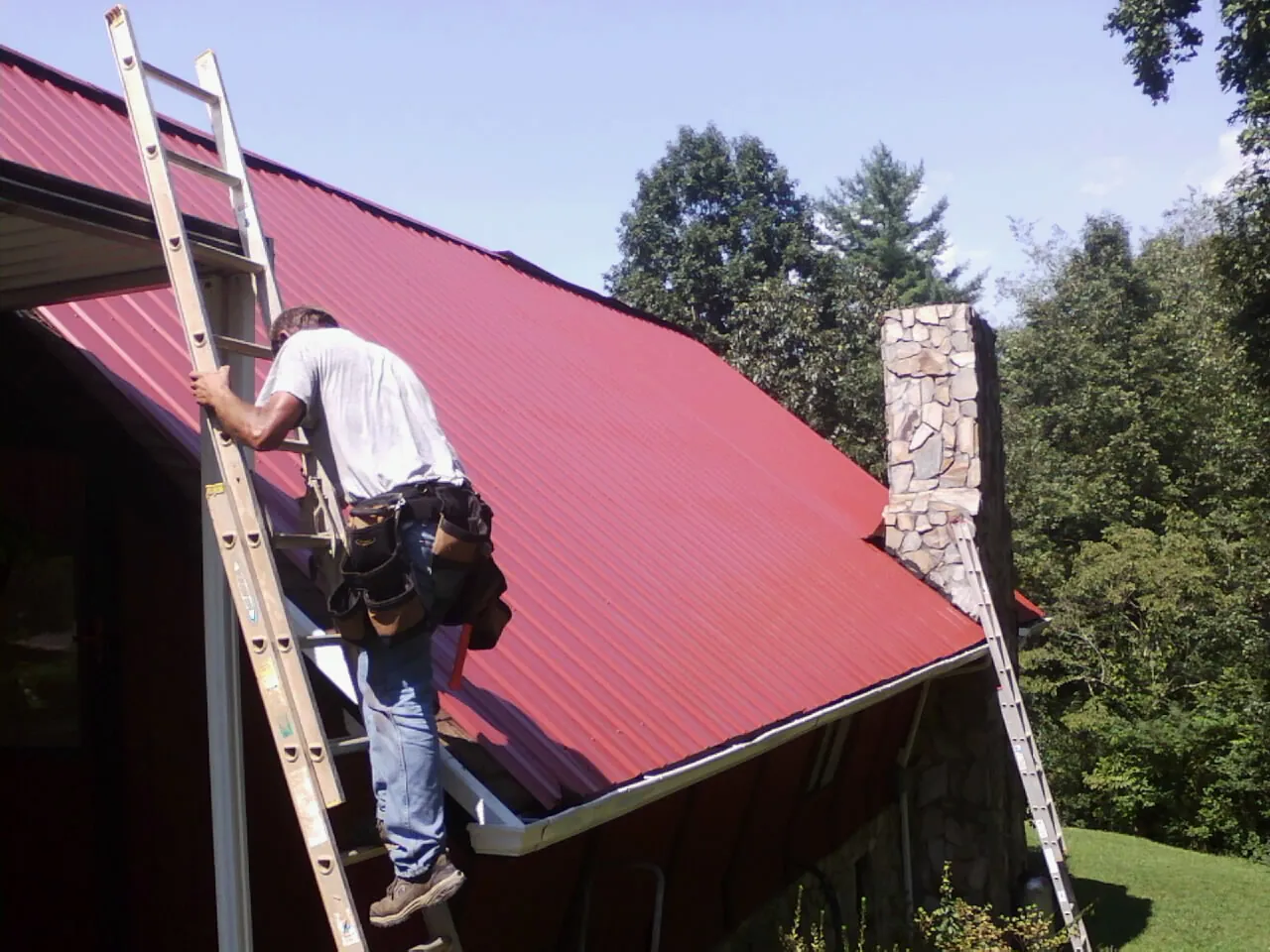 Roofer on a ladder beside a red metal roof and stone chimney — Soffit & Fascia Repair by Roofing Brothers in Shrewsbury